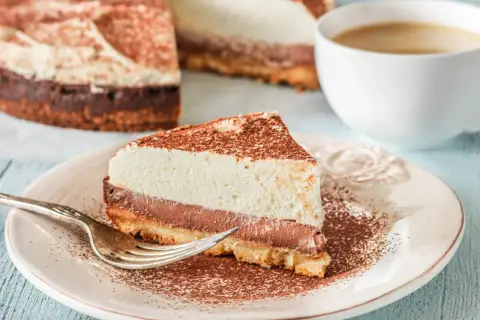 Pyszna tarta z mascarpone i czekoladą, posypana kakao, czeka na widelczyk. Obok filiżanka kawy.